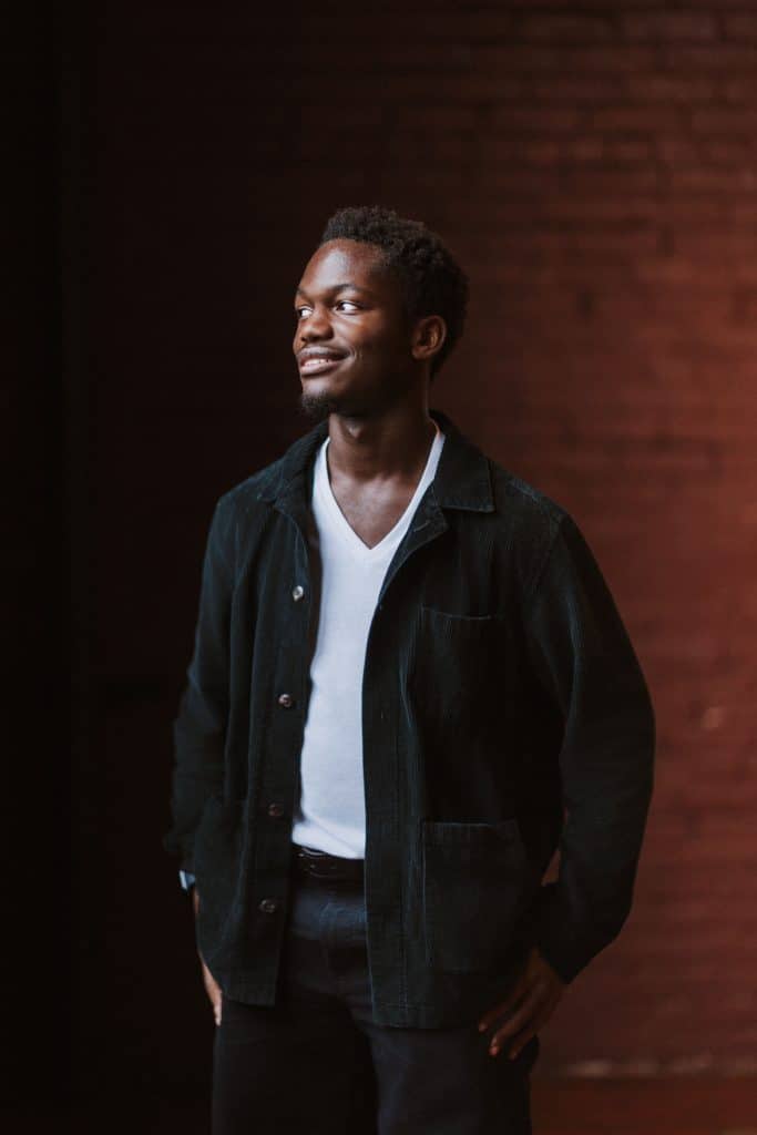 Senior portrait of a young man in a dark jacket and white t-shirt against a red brick wall