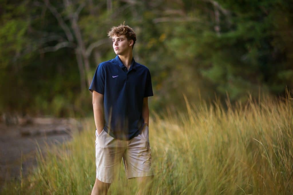 Senior portrait of a young man in a navy shirt and shorts standing in tall marsh grass