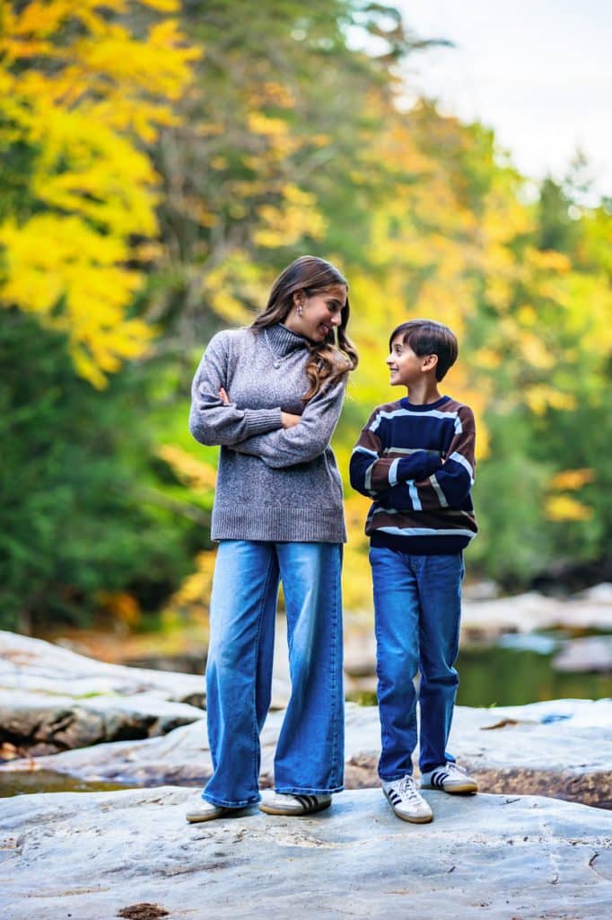 Woman and boy in sweaters and jeans standing together on riverside rocks beneath yellow fall foliage