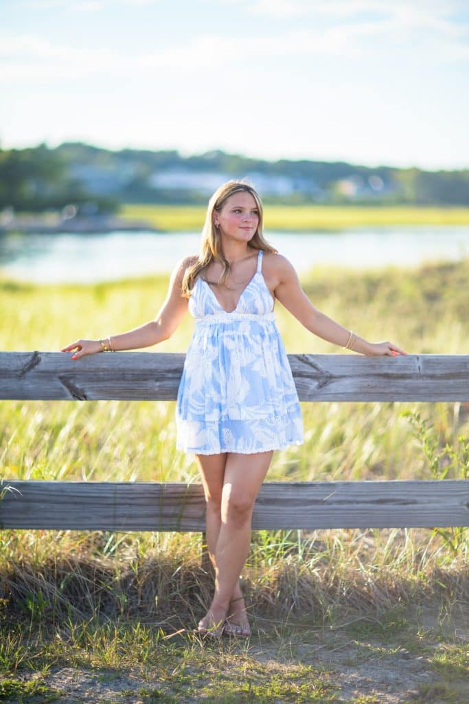 Senior portrait of a young woman in a light blue sundress leaning on a wooden fence in golden marsh light