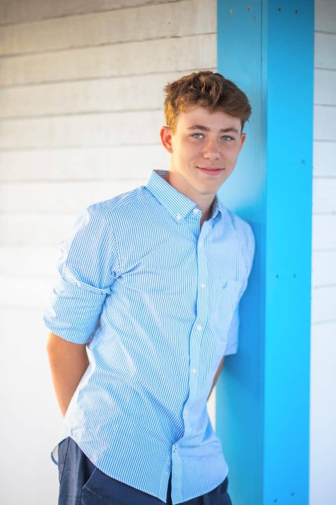 Senior portrait of a teenage boy in a blue striped shirt leaning against a bright blue door frame