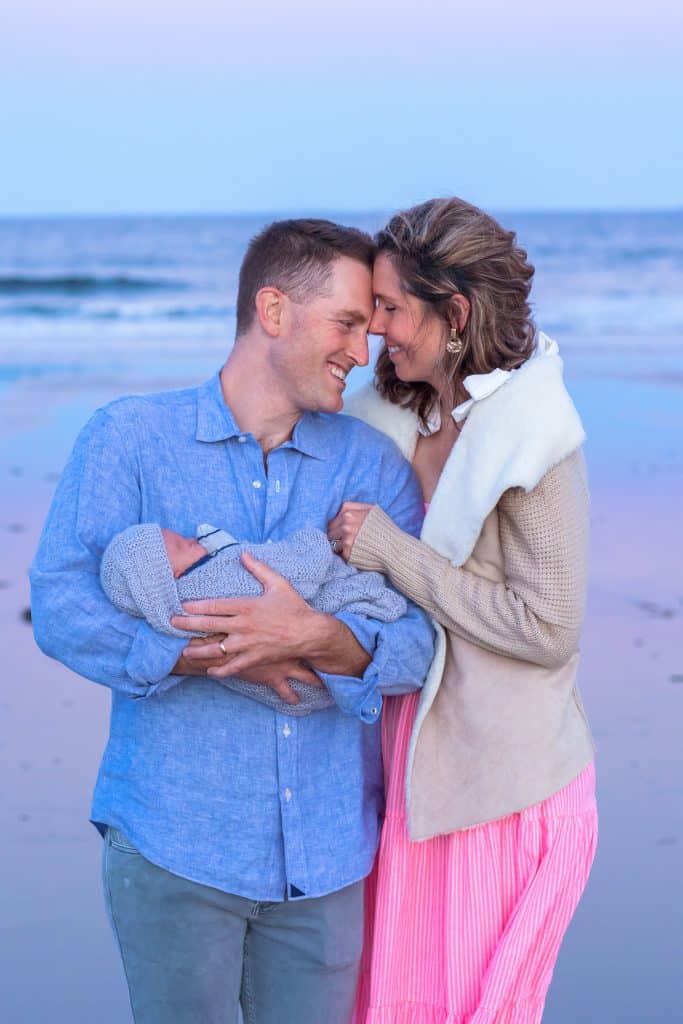 Couple embracing at the beach at dusk with their newborn baby wrapped in a blanket
