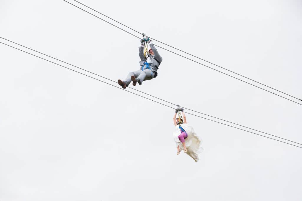 Two people ziplining side by side on parallel cables against a bright overcast sky