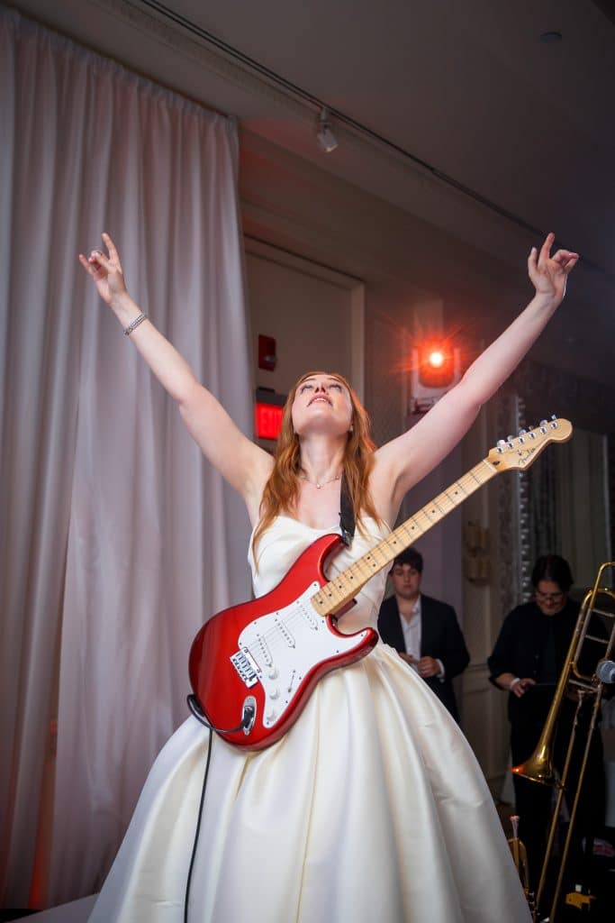 Bride in a ball gown raising her arms while holding a red electric guitar in front of a curtained window