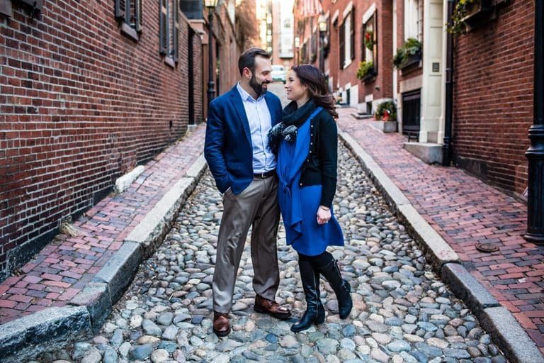 Engaged couple in blue coats standing on a cobblestone street lined with brick townhouses in Beacon Hill, Boston