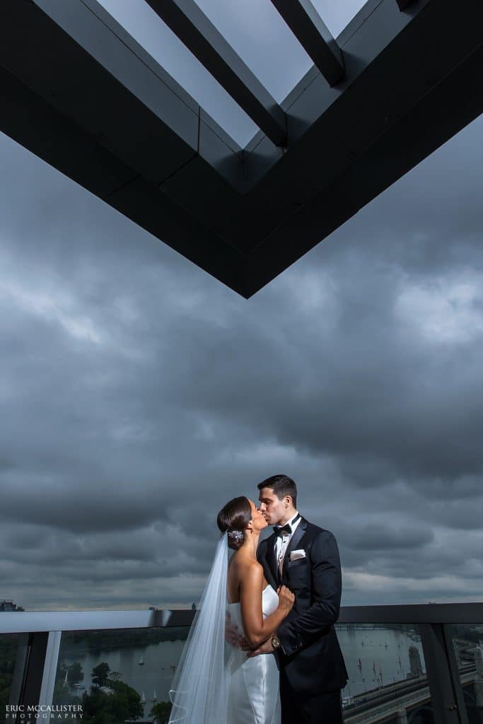 Bride and groom kissing on a hotel terrace beneath a dark storm sky and a modern angular roofline