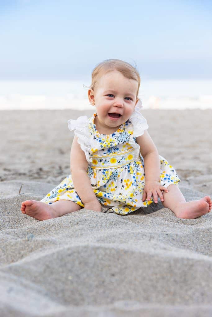 Baby girl in a yellow floral dress laughing while sitting on a sandy beach