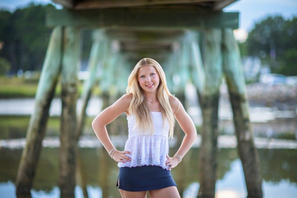 Senior portrait of a smiling young woman in a white top standing under a wooden pier with hands on hips